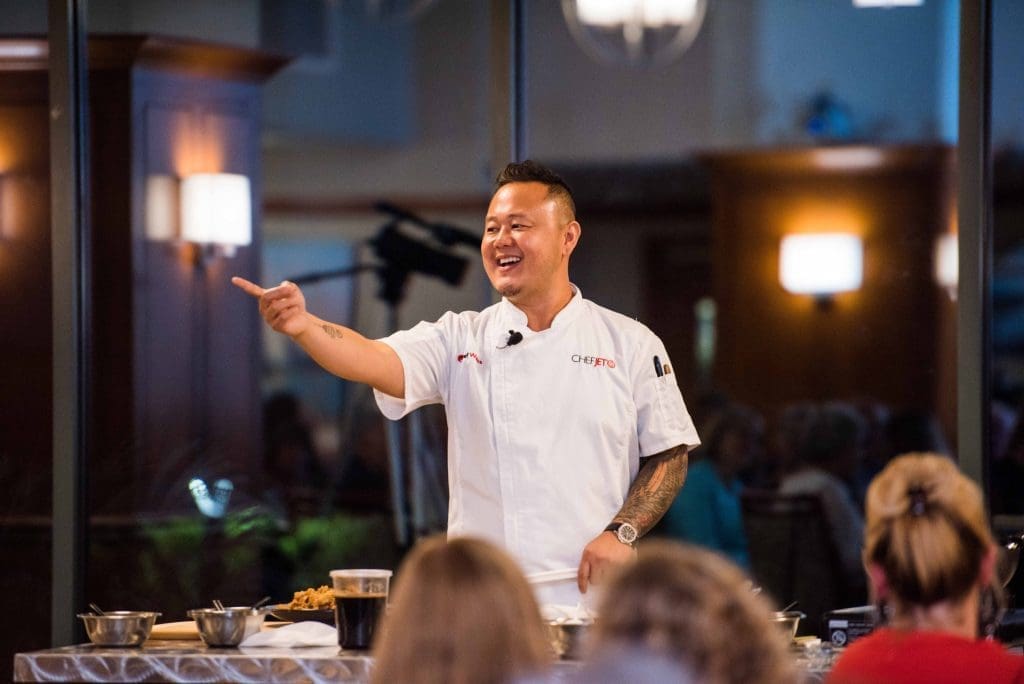 Chef Jet Tila in a white chef coat smiles and points while speaking to an audience at a cooking demonstration.