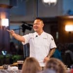 Chef Jet Tila in a white chef coat smiles and points while speaking to an audience at a cooking demonstration.