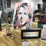 Painted portrait of a man with bold orange and blue strokes on a display table at the Kick Start Gallery, with "Hustle Harder" sign.