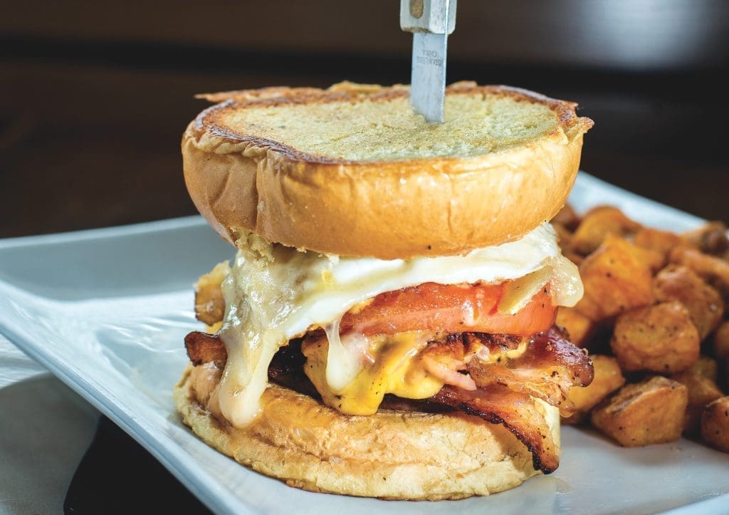 Stacked brunch sandwich with melted cheese, tomato, and bacon on a toasted bun, served with cubed breakfast potatoes.