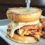 Stacked brunch sandwich with melted cheese, tomato, and bacon on a toasted bun, served with cubed breakfast potatoes.