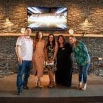 Five smiling adults pose on a raised stage in a bar with a stone accent wall and wall-mounted TV.