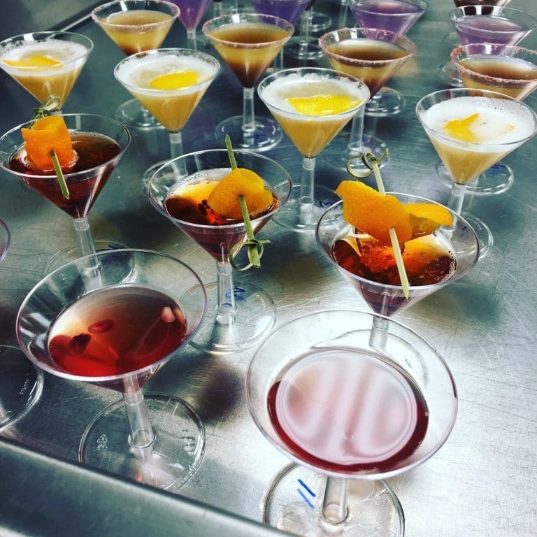 Rows of martini-style cocktails on a steel counter, some with frothy tops and orange peel garnishes, others deep red.