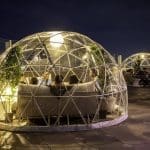 Guests sit inside illuminated clear geodesic dome enclosures on a rooftop terrace at night, with plants and sofas.