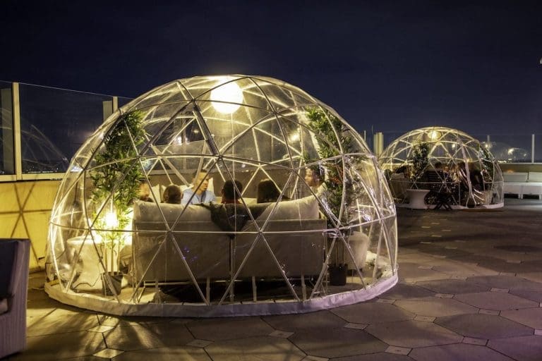 Guests sit inside illuminated clear geodesic dome enclosures on a rooftop terrace at night, with plants and sofas.
