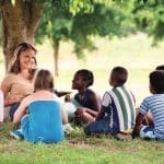 Children and education, young woman at work as educator reading book to boys and girls in park