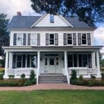 A white two-story farmhouse with a covered front porch, gray shutters, white columns, and a brick walkway.