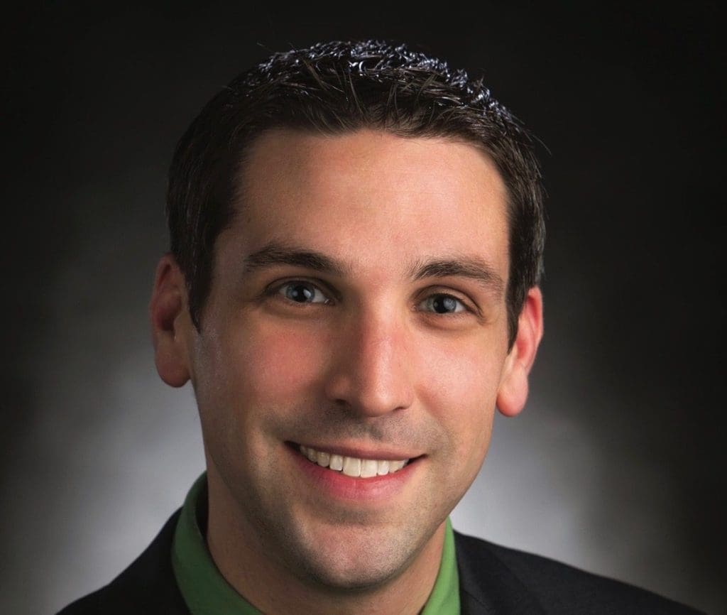 A smiling man with short dark hair wears a dark jacket and green collared shirt against a dark background.