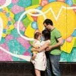 A family of three stands embracing in front of a colorful geometric mural with a large yellow sun motif.