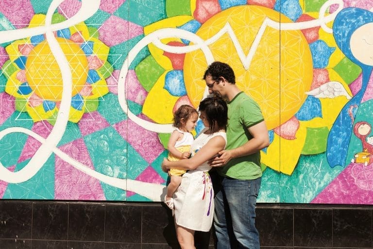 A family of three stands embracing in front of a colorful geometric mural with a large yellow sun motif.