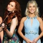 Four women in formal gowns stand against a dark backdrop, one playing a violin, smiling for the camera.