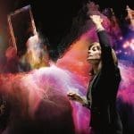 A conductor in a dark jacket raises one arm and holds a baton amid swirling pink and orange clouds and glowing music notes.