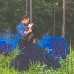 A couple in a forest embraces closely, the woman in a black lace-bodice gown amid billowing blue smoke.