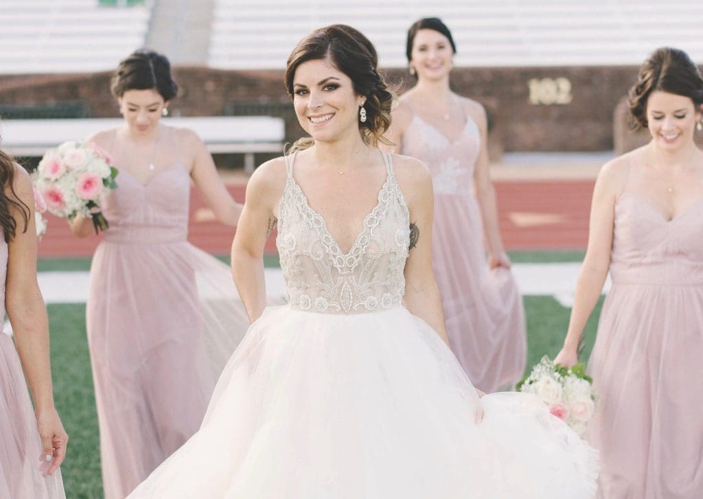 A smiling bride in a lace-bodice ball gown walks across a football field with bridesmaids in blush dresses behind her.