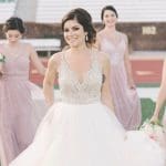 A smiling bride in a lace-bodice ball gown walks across a football field with bridesmaids in blush dresses behind her.