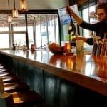 A bartender in glasses mixes drinks at a long wood bar lined with teal-backed stools and metal shakers.