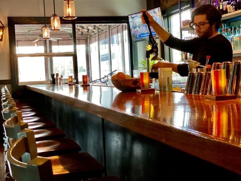 A bartender in glasses mixes drinks at a long wood bar lined with teal-backed stools and metal shakers.