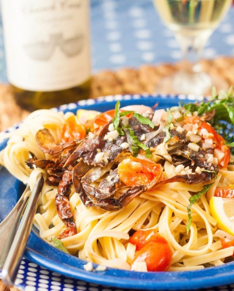 Fettuccine topped with sautéed soft-shell crabs, cherry tomatoes, garlic, and basil on a blue plate beside a bottle of white wine.