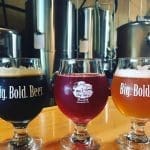Three tulip glasses of dark, red, and amber beer in front of steel brewing tanks, with "Big. Bold. Beer." and "Big Ugly Brewing" logos.