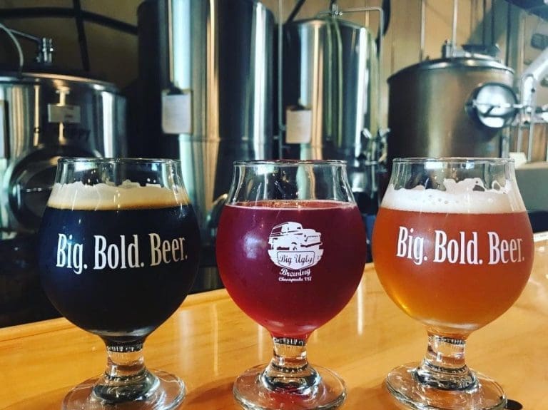 Three tulip glasses of dark, red, and amber beer in front of steel brewing tanks, with "Big. Bold. Beer." and "Big Ugly Brewing" logos.