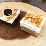 Two square dessert bars with toasted meringue tops on a wooden slice, one topped with a chocolate sandwich cookie.