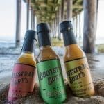 Three bottles of Bootstrap Burt's hot sauce stand in wet sand beneath a wooden pier over the ocean.