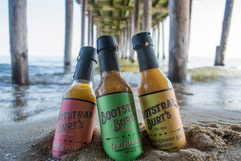 Three bottles of Bootstrap Burt's hot sauce stand in wet sand beneath a wooden pier over the ocean.