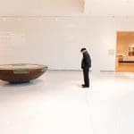 A man in a dark cap stands by a low bronze bowl sculpture in a white gallery marked Gallery M.