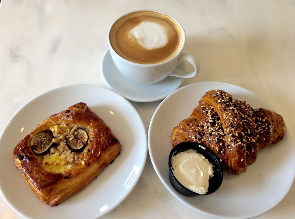 A latte with foam art beside a fig and nut pastry and a sesame-topped croissant with cream cheese on marble.