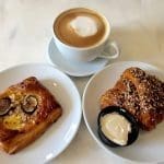 A latte with foam art beside a fig and nut pastry and a sesame-topped croissant with cream cheese on marble.