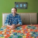 A smiling man in a blue plaid shirt sits at a table with an orange floral tablecloth under green walls.