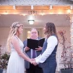 A bride in a flower crown and lace dress holds hands with her partner before an officiant on a string-lit porch.