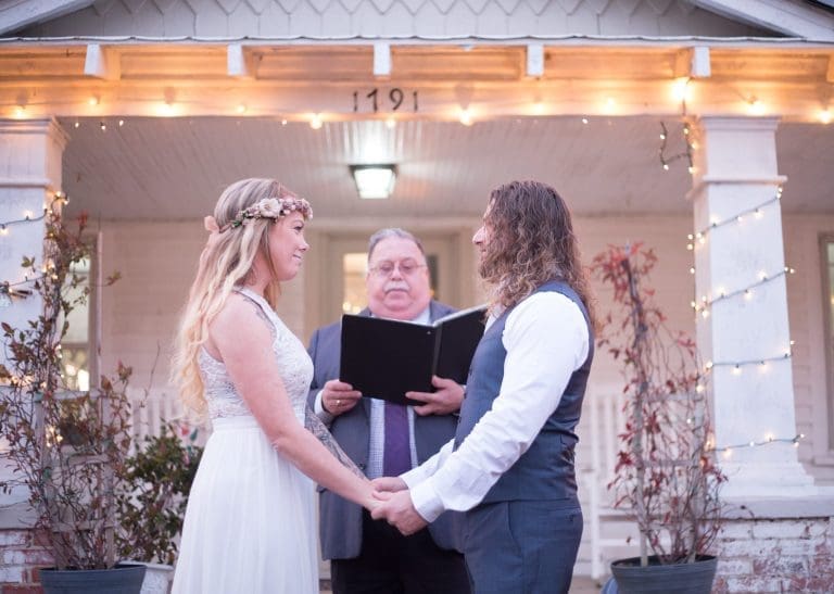 A bride in a flower crown and lace dress holds hands with her partner before an officiant on a string-lit porch.