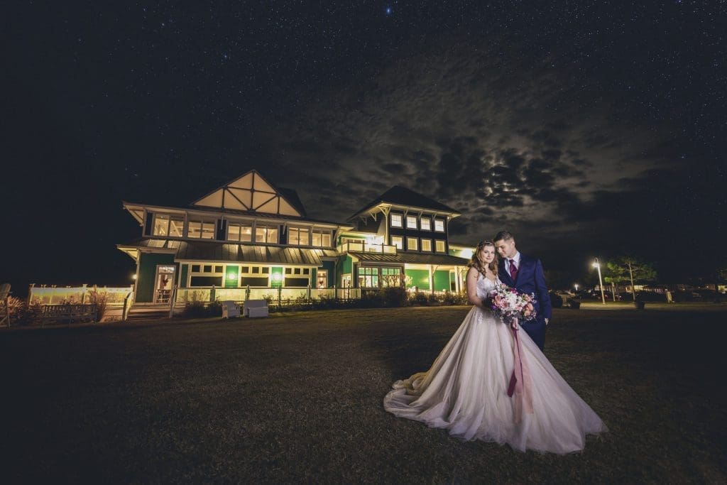 A bride in a ballgown and a groom in a navy suit pose before a lit green Victorian-style clubhouse under a starry night sky.