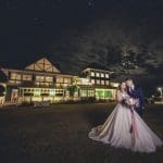 A bride in a ballgown and a groom in a navy suit pose before a lit green Victorian-style clubhouse under a starry night sky.