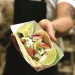 A person holds a paper tray with a tortilla taco topped with pulled meat, slaw, tomato, onion, cream and lime wedges.