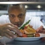 A chef in a white jacket leans over a plate of seared salmon, asparagus, tomato salsa, and cherry tomatoes.