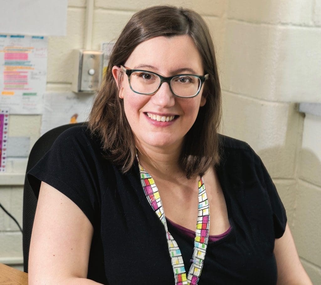 A smiling woman with dark-framed glasses and a colorful lanyard sits at a desk against a cinderblock wall.