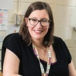 A smiling woman with dark-framed glasses and a colorful lanyard sits at a desk against a cinderblock wall.