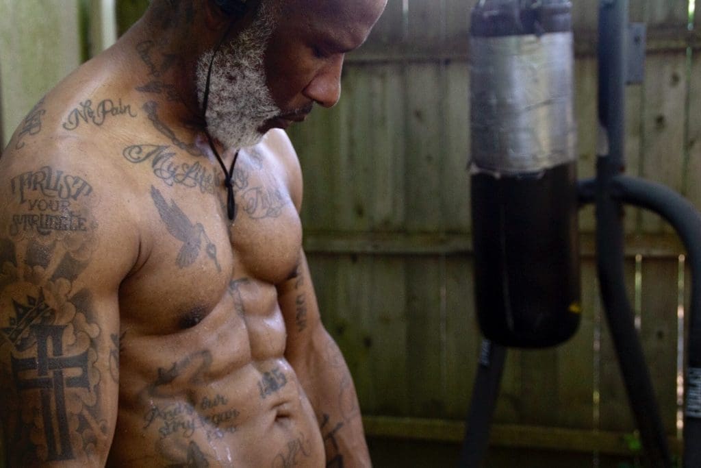 A shirtless, tattooed man with a gray beard looks down beside a heavy bag in a backyard.